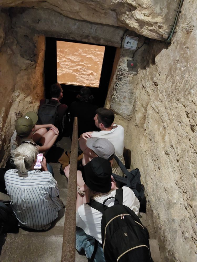 Eine Gruppe von Exkursionsteilnehmenden befindet sich in einem schmalen, in den Fels gehauenen Gang. Sie sitzen auf einer Treppe und blicken durch eine Glaswand in einen beleuchteten Raum.