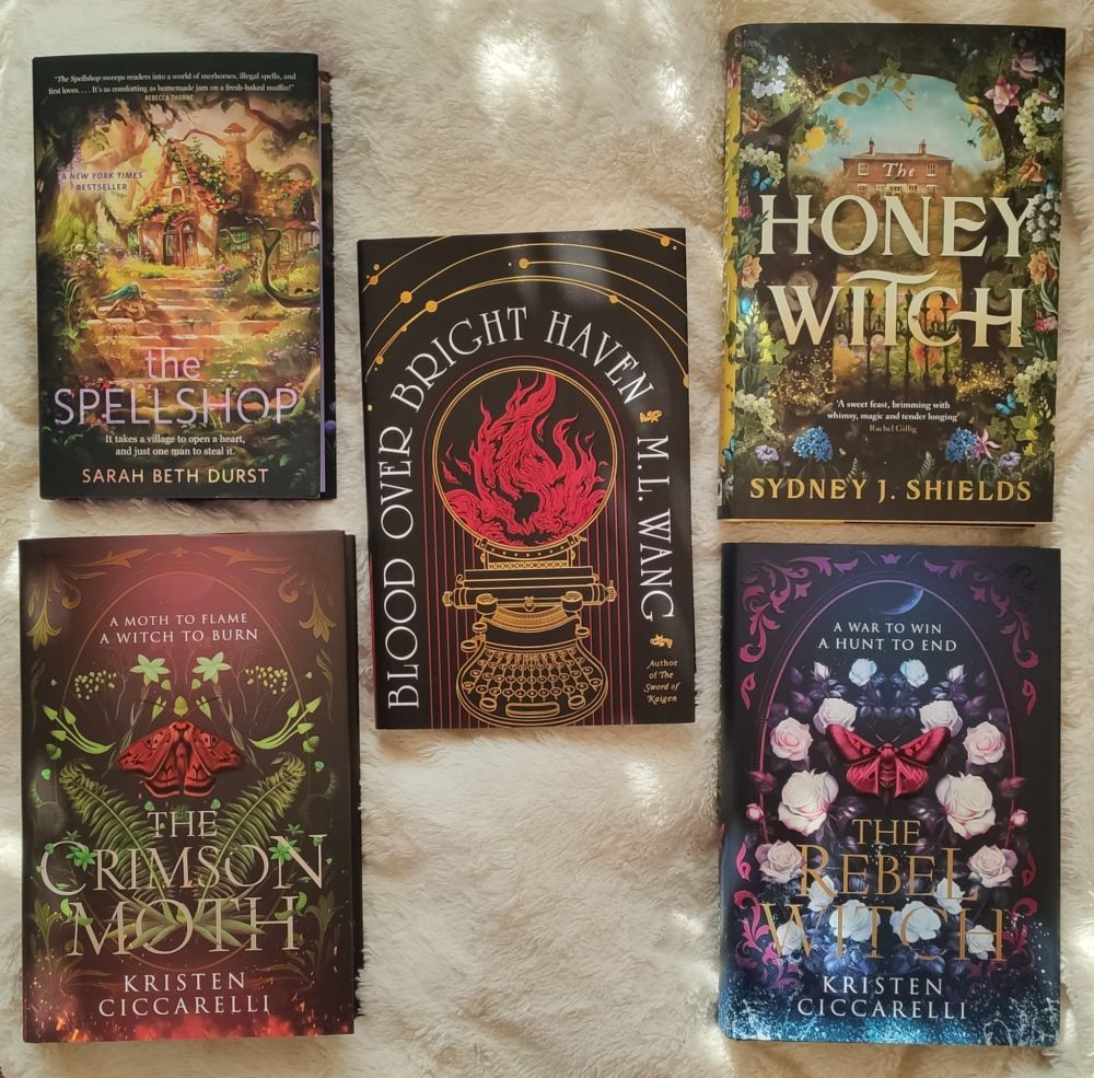 Five books lying on a soft, fluffy white pillow. Those books are The Spellshop by Sarah Beth Durst, Blood Over Bright Haven by M.L. Wang, The Honey Witch by Sydney J. Shields, The Crimson Moth and The Rebel Witch by Kristen Ciccarelli.