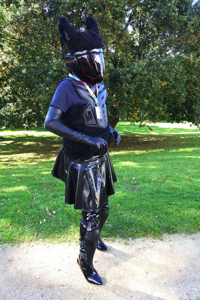 Fursuit photo: Quirk the protogen wearing latex opera gloves and skirt next to the lawns of the Pawsome venue.  He also is wearing long high heeled boots with paintball armour over them.