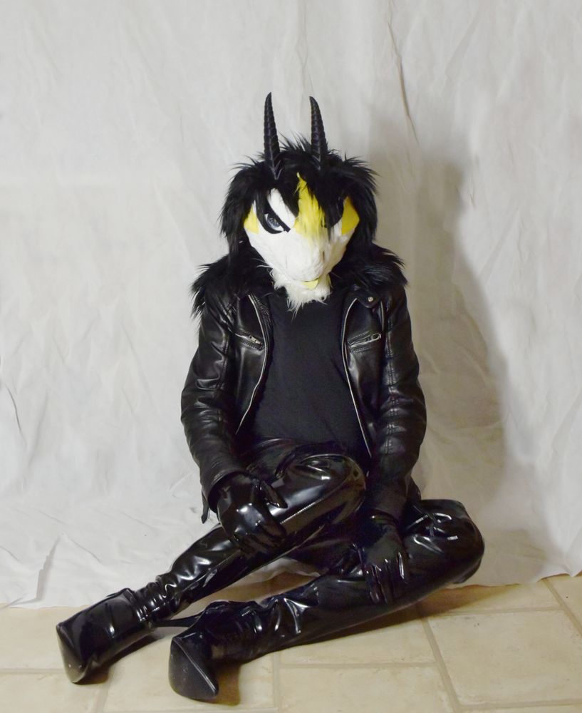 Fursuit photo of an anthro dragon wearing a black leather jacket, PVC gloves, long PVC boots with stiletto heels and rubber jeans.
He is sitting on the floor in front of a white photographic backdrop