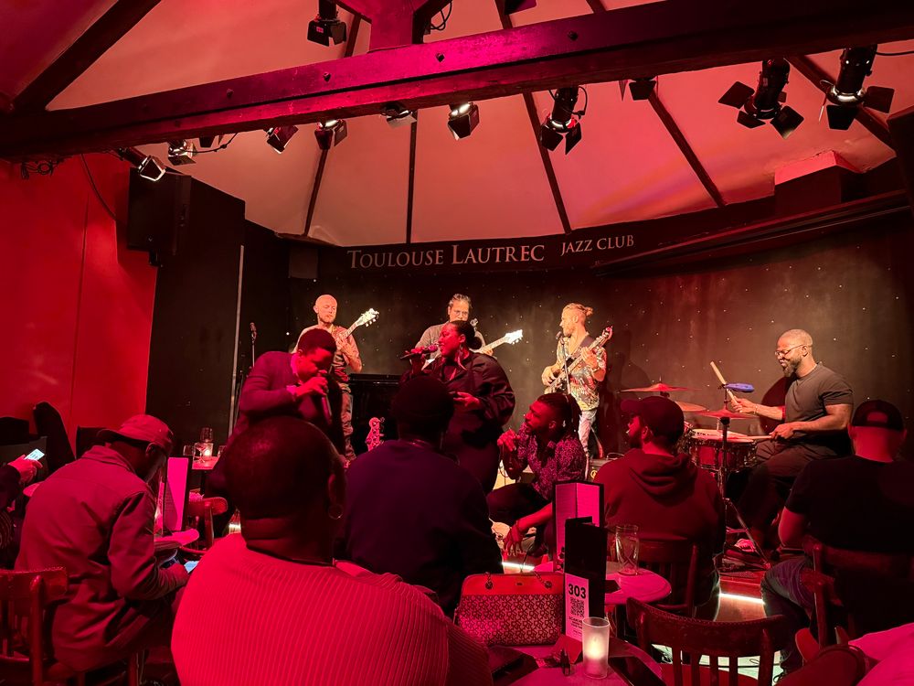 A group of cool looking musicians up on a small stage in a jazz club. There are starry lights on the wall. 