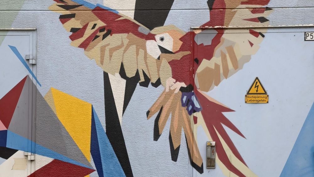 Nahaufnahme eines komplett mit Graffiti übermalten Stromkastens. Das Graffiti im kubischen Stil zeigt einen in Beige, Rot und Schwarz gemalten Adler, der seine Flügel aufschwingt. Dass es sich um einen Stromkasten handelt, ist an einem gelben Warndreieckschild mit einem Blitz und der Unterschrift "Hochspannung! Lebensgefahr" erkennbar.