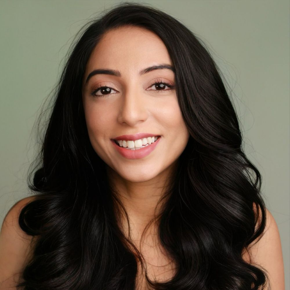 Headshot of Sneya Rajani, a South Asian woman with wavy black hair wearing a grey tank top, smiling against a soft green backdrop.