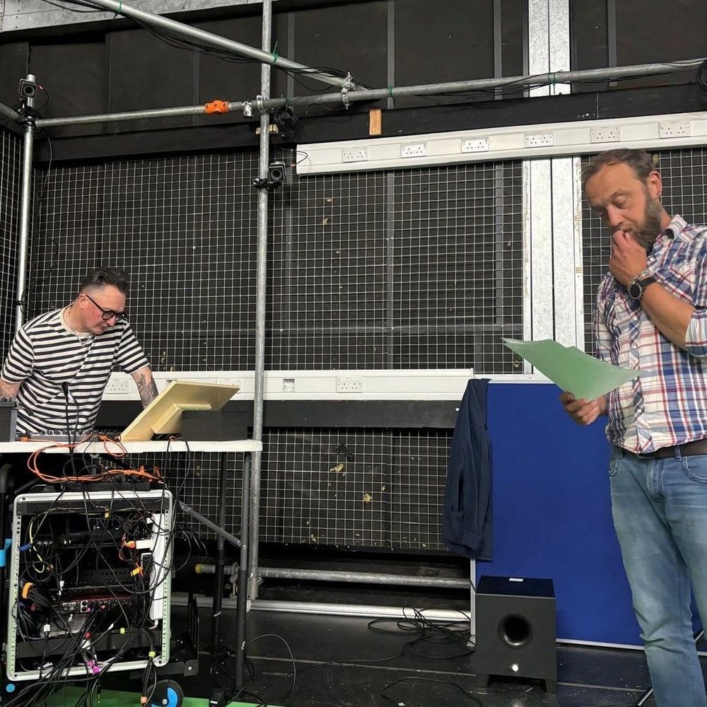 Two men, one bearded man wearing a checked shirt leaning on a table writing with another seated wearing a baseball cap, headphones and striped t-shirt against a green screen background.