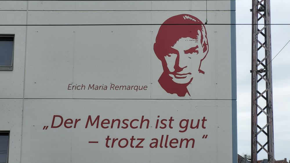 Das Bild zeigt eine Hauswand mit einem stilisierten Porträt von Erich Maria Remarque. Links neben dem Porträt steht sein Name in roter Schrift. Darunter befindet sich ein Zitat das lautet:

"Der Mensch ist gut – trotz allem".