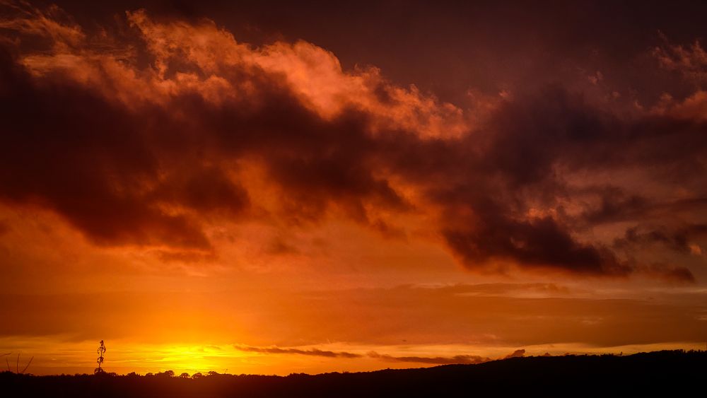 Dieses Bild zeigt einen intensiven Sonnenuntergang mit einem Himmel, der von dichten, dramatischen Wolken dominiert wird. Die Wolken sind in tiefen Orange-, Rot- und Brauntönen gefärbt, während die Sonne knapp über dem Horizont steht und ein warmes, goldenes Licht ausstrahlt. Die Stimmung wirkt fast düster und mystisch, besonders durch die dunklen Silhouetten der Wolken. Am unteren Rand des Bildes sind die Umrisse einer Landschaft oder von Hügeln zu erkennen, die sich dunkel gegen den leuchtenden Himmel abheben.