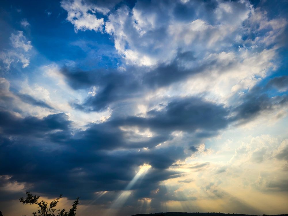 Eine dramatische Wolkenformationen in verschiedenen Grautönen, die sich mit leuchtend blauen Himmelsflecken vermischen. Sonnenstrahlen brechen durch eine Lücke in den dunkleren Wolken am unteren Bildrand und erzeugen helle, vertikale Lichtstrahlen, die den Vordergrund beleuchten. Am Horizont ist eine dunkle Silhouette einer Landschaft oder von Bäumen zu erkennen
