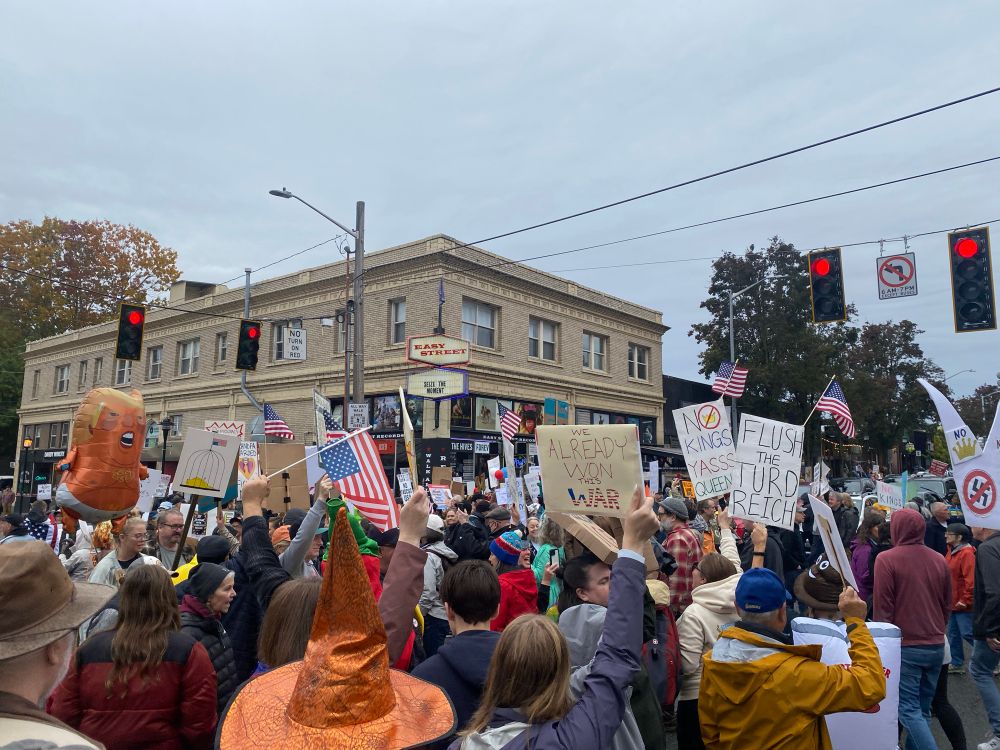 Hundreds of people fill the streets in front of  a two-story brick building with a red neon bordered sign reading EASY STREET.  Below that sign is a marquee sign reading SEIZE THE MOMENT. People wear festive hats and costumes including an orange witch’s hat and a red chili pepper costume, wave American flags and an angry baby Trump in a diaper balloon, and carry signs reading “NO KINGS”, “WE ALREADY WON THIS WAR”, “NO KINGS, YASS QUEEN!” a swastika with a red slash through it, and “FLUSH THE TURD REICH”. 
US demonstrators celebrating their civil rights on NO KINGS DAY, October 18, 2025, in front of Easy Street Records, Alaska Junction, intersection of California Ave SW and SW Alaska Street, West Seattle, Washington state, USA.