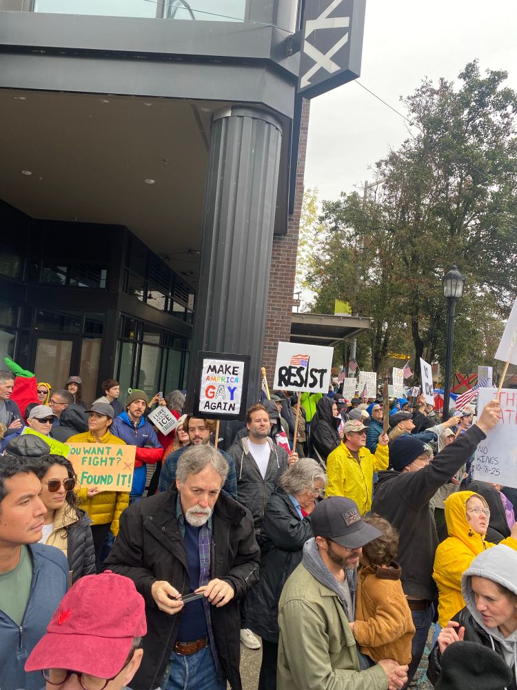 People crowd a street corner waving American flags and signs reading “RESIST”  (with an upside down American flag), “MAKE AMERICA GAY AGAIN” (with AMERICA written in rainbow colors), “YOU WANT A FIGHT? YOU FOUND IT!”, “I LIKE MY ICE CRUSHED”, “FLUSH THE TURD REICH”, and “NO KINGS”. West Seattle, Washington. NO KINGS Day. October 18, 2025.