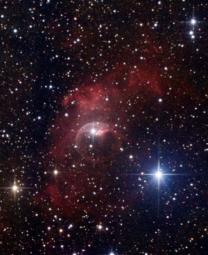 A red cloud surrounded by many hundreds of stars. There are several very bright blue, white, and yellow stars with prominent diffraction spikes. Near the center of the red cloud is a somewhat fainter pinkish ring, somewhat like a soap bubble.