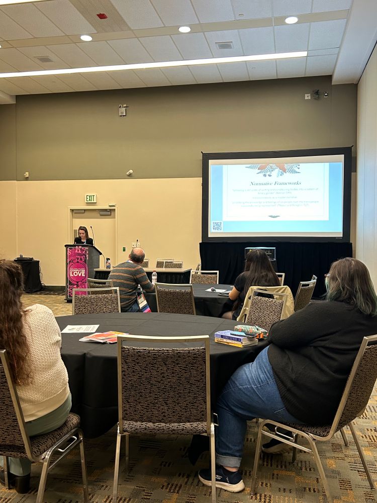 A speaker addresses a room of conference attendees. A large screen showcases slides on the right side of the room.
