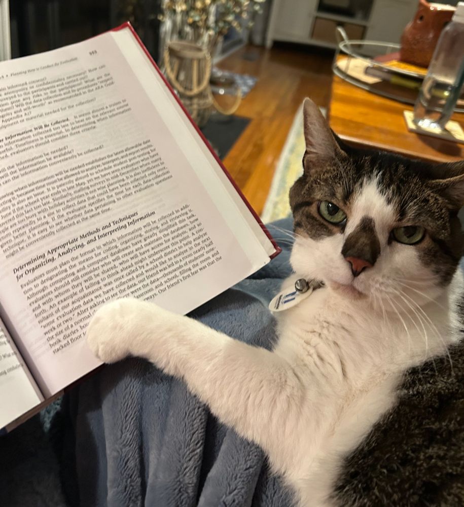 A cat rests her paw on a large textbook and looks smugly at the camera, almost like she has a secret.