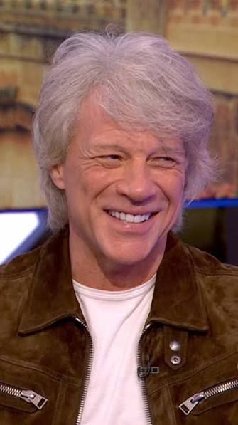 Wispy white haired man with crinkled features smiles wearing a white t-shirt and brown suede jacket. 