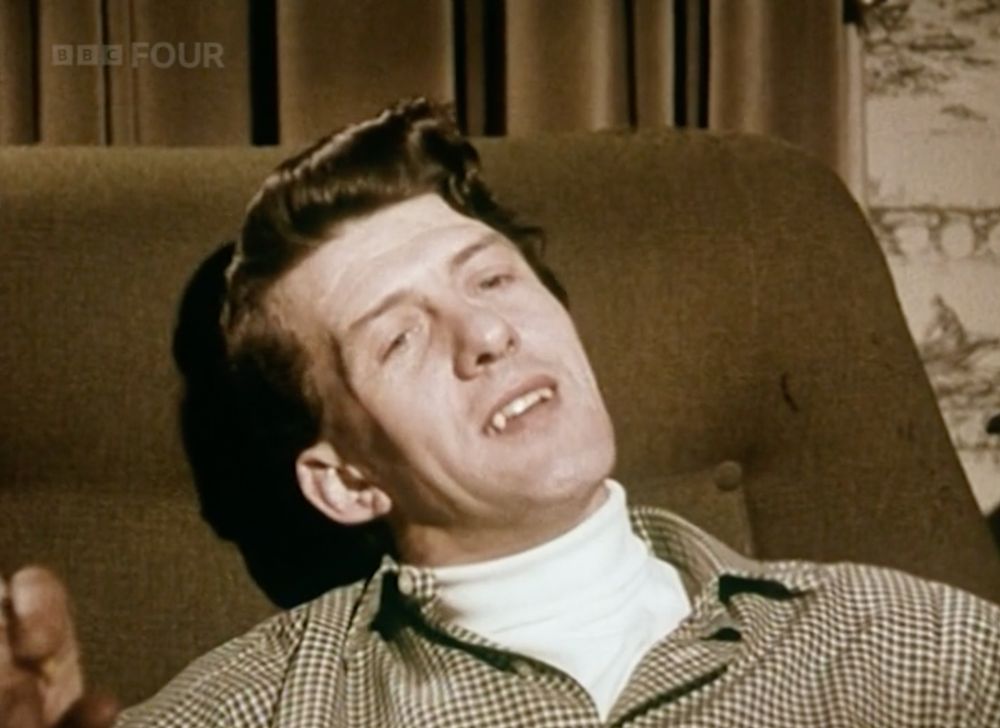 Still image from 1969 BBC documentary. Man sitting in chair with excessively oiled hair with mouth slightly open revealing large pointy canine teeth like Dracula.