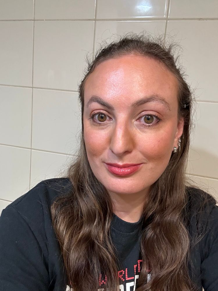 A woman with brown hair and the sides pulled back. She has a plum smokey eye and a dewy deep pinkish red lip oil on. 