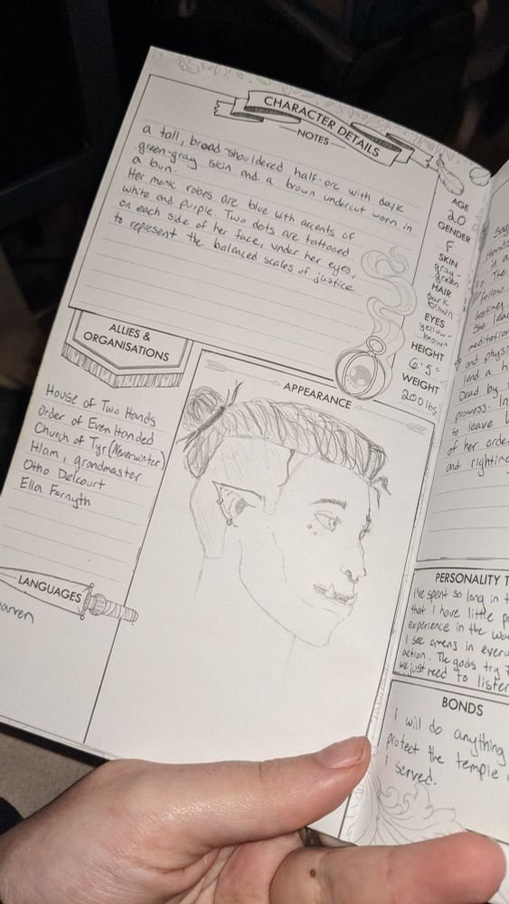 A photo of a D&D 5E character journal showing a rough sketch of a half-orc woman with her hair in a bun and sides of her head shaved. She has two dots tattooed under her visible eye and a septum ring. Listed description reads: "a tall, broad-shouldered half orc with dark green-gray skin and a brown undercut worn in a bun. Her monk robes are blue with accents of white and purple. Two dots are tattooed on each side of her face, under her eyes, to represent the balanced scales of justice."
