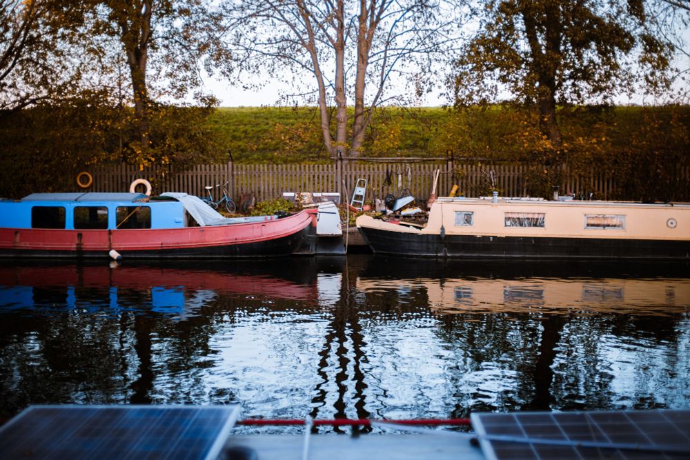 A peaceful canal scene with two narrowboats moored side by side - one red and blue, the other cream - with their reflections rippling in the water. Behind them is a wooden fence, scattered items like a bicycle and tools, and tall autumn trees with sparse leaves.