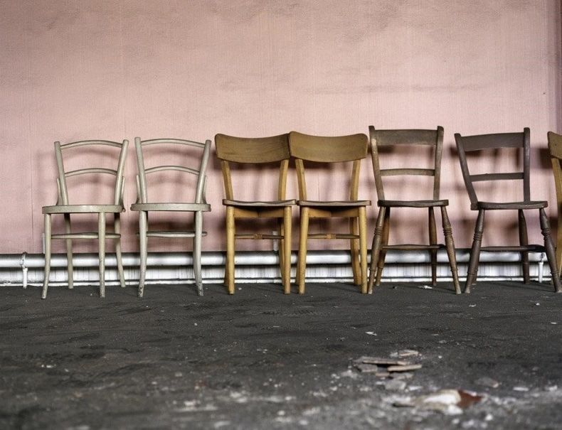 Image: a photo of seven wooden chairs against a somewhat grubby pale-pink wall; we are viewing them ‘face-one’ at seat level, and the chairs are ranged from left to right across the photo, sitting on a dark-grey floor of some sort, perhaps carpet; we also see white pipes behind the chairs, just above the floor, behind the chairs; the first two chairs are white-ish, the next two yellowish wood, the next two grey-brown wood, the last one like the third and fourth ones, but severely cropped off because at the right edge of the photo; something has been smashed on the floor near the camera • “Ethna Rose O’Regan, Sinead McCann in collaboration with Dr Louise Brangan  Magdalene Laundries were once vast islands of abandonment marking our Irish social landscape. They are closed now, but their legacies linger. When the gates were locked for the last time in 1996, it was with a whimper not a bang. The secrets were sealed off, and the nation moved on, or so it seemed. But this recent…”