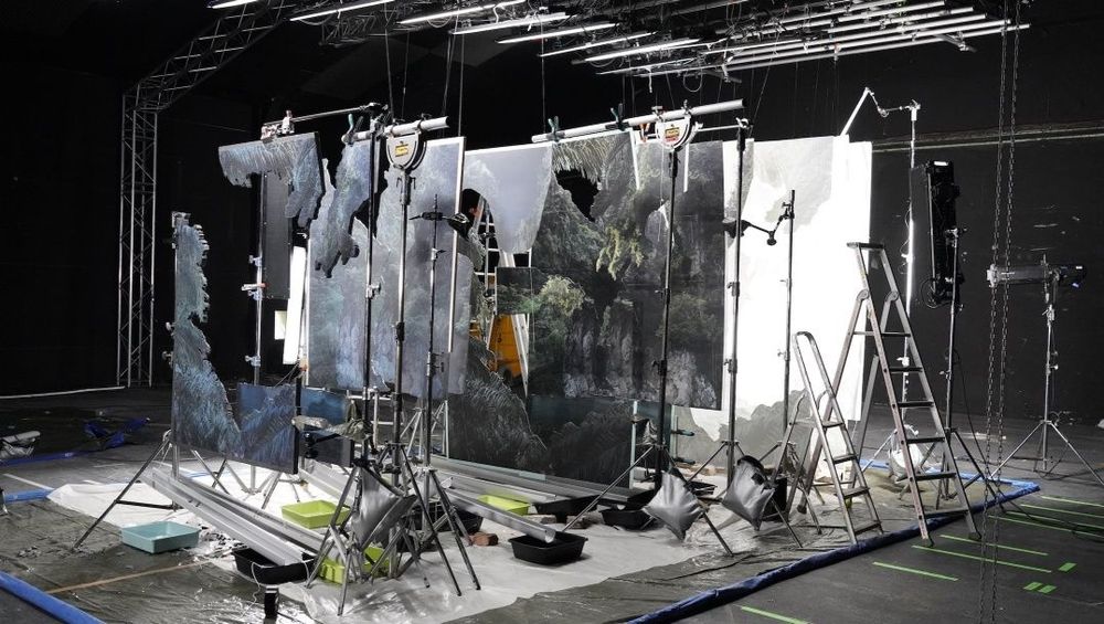 Image: Noémie Goudal, behind the scenes of Supra Strata, 2024, courtesy of the artist and Edel Assanti, London | photo showing a series of ‘canvases’ – pieces of material, often with large parts missing – hanging down from supports, one behind the other, such that someone looking from in front of the arrangement would see a series of layers, most pierced to reveal a further background behind; lighting is from above, and the space itself is painted black • “Noémie Goudal constructs illusionistic interventions in the landscape, captured through photography, film and sound. Deploying scenography and staging devices, she expands photography beyond its conventional parameters into immersive installations, informed by her interest in paleoclimatology. While human temporality does not align with the deep time of geological processes, she seeks to visualise the effort that goes into making the landscape.    Created for the Prix Marcel Duchamp, Supra Strata (2024) is a…”