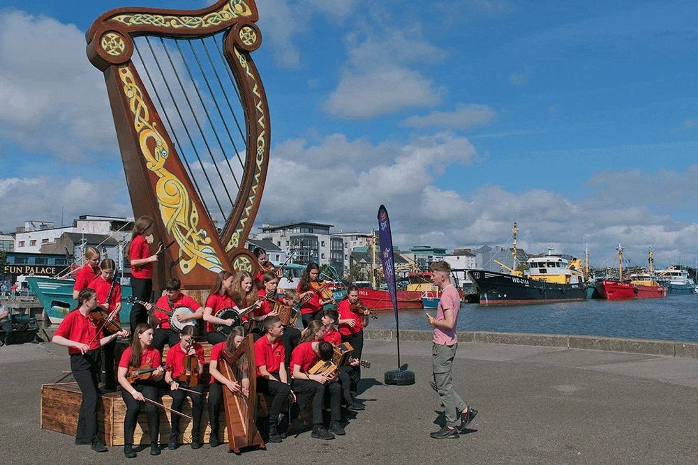 Image: the photo, taken on a waterfront pier, shows a group of young (probably teen) musicians in red tops and dark trousers holding various instruments as they attend to their director, apparently equally young, standing in front of them in a pink t-shirt and grey trousers; towering over them is a Celtic harp with Celtic design; in the background we see buildings and ships; it's sunny with a bit of cloud • “Wexford Arts Centre and Wexford Camera Club are proud to present In the Moment: Capturing the Fleadh 2024, a compelling group photography exhibition that celebrates the vibrant energy and cultural richness of last year’s Fleadh Cheoil. This exhibition showcases the spirit of the festival through the lens of twelve talented photographers, all members of the Wexford Camera Club. The photographers include Darren Arkins, Bart Bulla, Kevin Cadogan, Miriam Furlong, Aoife Kavanagh, Donna Kehoe, Lucy Leonel, Alan Mahon, Ann McCafferty, Ian Moriarty, John Mullins, and Marie Phelan…”