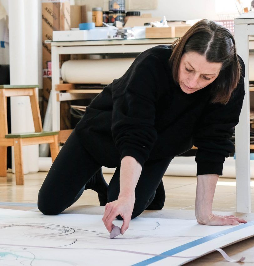 image: a photo, presumably of Órla Bates, on hands and knees in black top and jeans, drawing on what may be paper or canvas that is lying on the floor; the shot is from in front of the artist, who is looking down at the mark being made by what may be a chunk of pigment of some sort, purplish; behind we seem to see an artist’s studio, and possibly very large rolls of paper • “A solo exhibition by Órla Bates featuring drawings made in collaboration with artist Joanna Leah • Curated by Ann Mulrooney    In 2023, Órla Bates was invited to take part in the MAKE/curate programme, a partnership initiative between Wexford Arts Centre, the Arts Department of Wexford County Council, and Blackbird Cultur-Lab. The aim of the programme is to provide artists working regionally with an opportunity to work with a national curator. Over the course of two years, Orla worked with Curator Ann Mulrooney towards her solo exhibition at Wexford Arts Centre. Distinct from the curatorial support, mentorship was…”