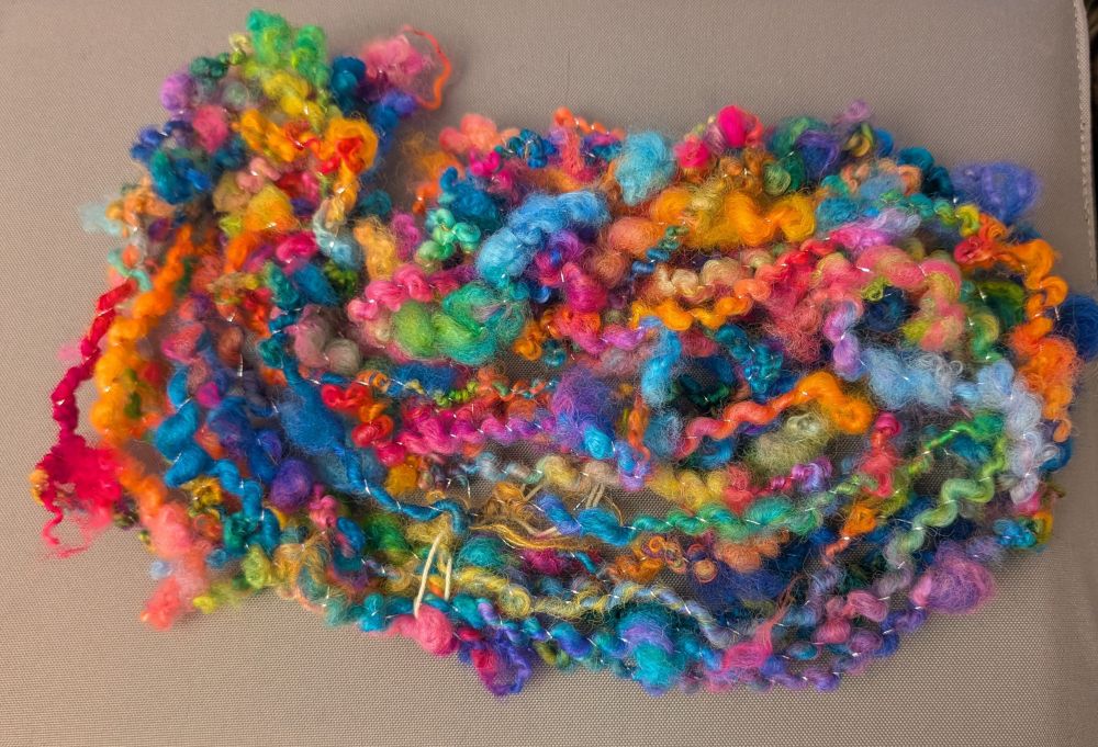A skein of textured fluffy handspun yarn laid on a grey mat. It's very bright rainbow locks in no particular order spiral plied with silver thread. The ends of the locks peek out here and there adding to the texture. 