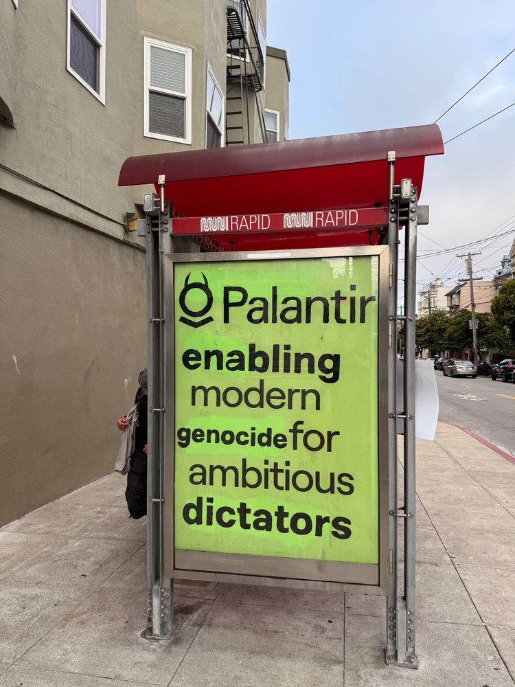 A San Francisco Muni bus shelter with its advertising space taken over by a bright green poster: "Palantir: Enabling modern genocide for ambitious dictators"  The Palantir logo has had devil's horns added.