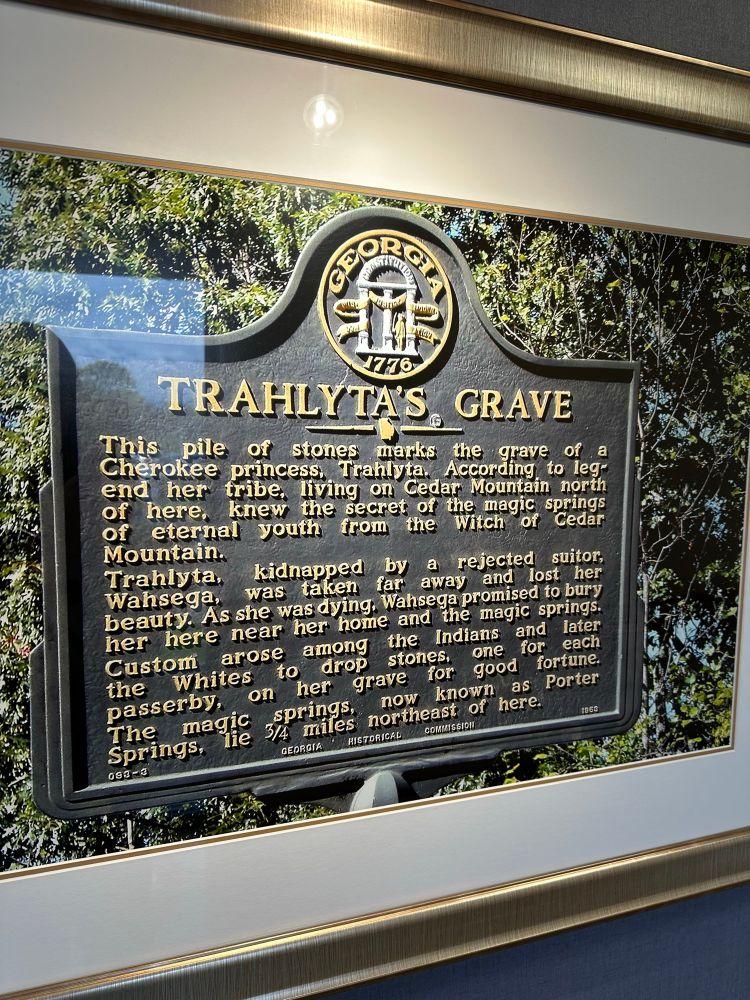 Photo of a historical marker. Reads:

TRAHLYTA'S GRAVE
This pile of stones marks the grave of a Cherokee princess, Trahlyta. According to legend her tribe, living on Cedar Mountain north of here, knew the secret of the magic springs of eternal youth from the Witch of Cedar Mountain.
Trahlyta, kidnapped by a rejected suitor, Wahsega, was taken far away and lost her
beauty. As she was dying. Wahsega promised to bury her here near her home and the magic springs. Custom arose among the Indians and later the Whites to drop stones., one for each passerby, on her grave for good fortune. The magic springs, now known as Porter Springs, lie 3/4 miles northeast of here.