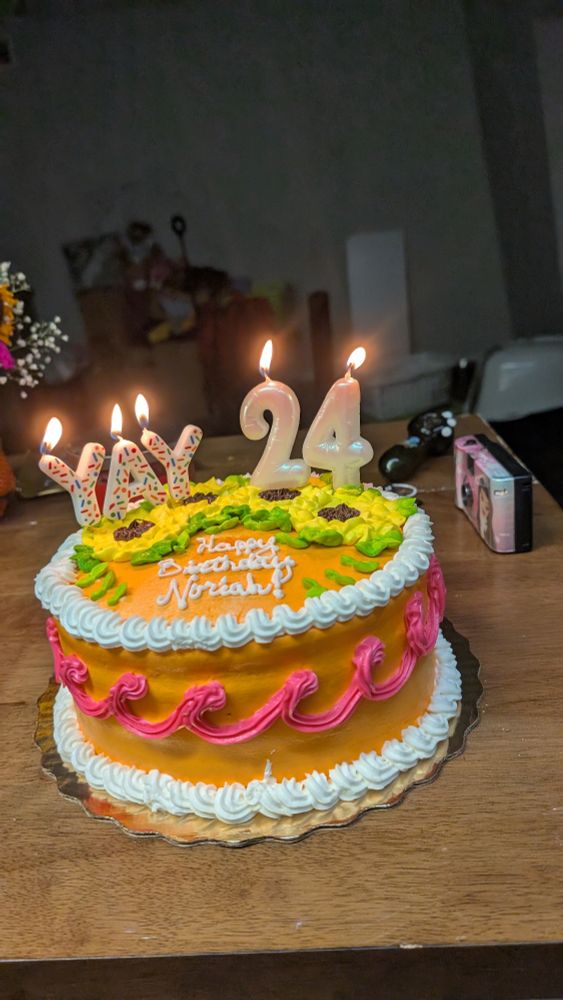 am orange cake with sunflowers on it that says "happy birthday Noriah!' and candles that spell out "YAY" and "24". there is an absolute mess in the background.