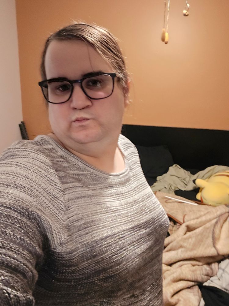 A picture of a oale trans women named ashe taking a selfie.

She has her long black hair tied back and has black framed glasses.

She is wearing a sweater that slowly changes from white to a grray grafian and sparsckles a little.

Ahe is making a pounty kissy lile face with her lips