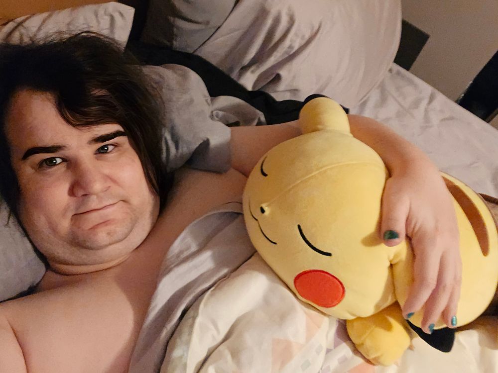 A picture of me ashe a pale white trans women lying in bed with blankets on snuggling my giant sleepy pikachu plush 