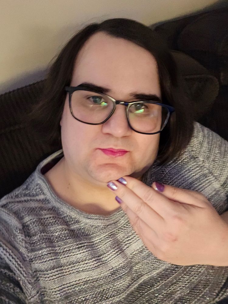 A picture of yours truely a pale white trans girl with long black hair and pink lipstick and glasses and finger nails painted cosmic purple.

She is wearing her sweater ehich is a gradiant of white and gray 