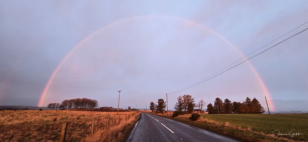 A road disappears into the distance towards a rainbow forming a dome over the rural landscape