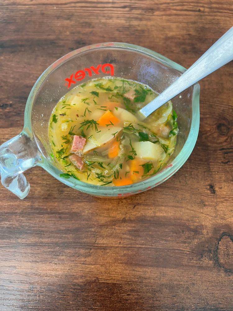 A Pyrex measuring cup of soup with carrots, potatoes, turkey sausage and herbs visible is on a wooden table with a spoon in it. 