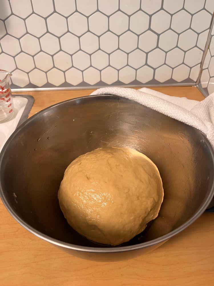 A yellow round of dough sits in a silver bowl with a towel partially draped over it 