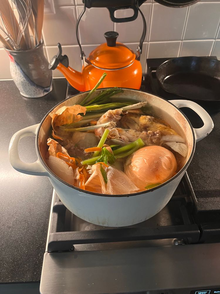 A white pot sits on a stovetop with various onions and celery and chicken bones 