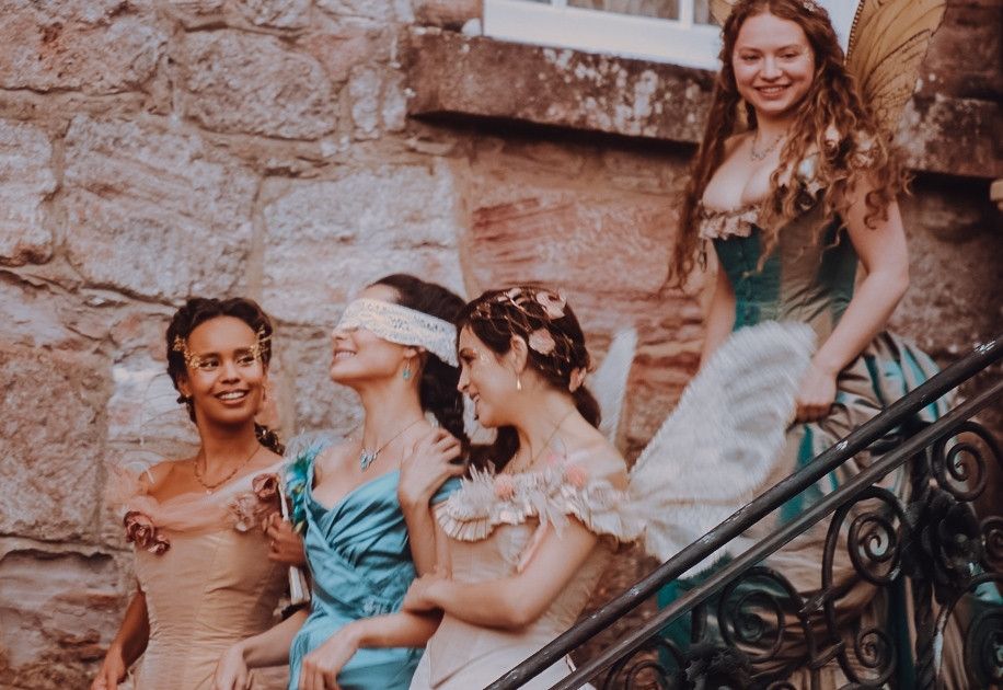 Meninas em roupas de epoca com vestidos longos descendo uma escadaria durante o dia com asas de fadas de costas para uma grande janela em tons pastéis de azul, rosa e laranja 