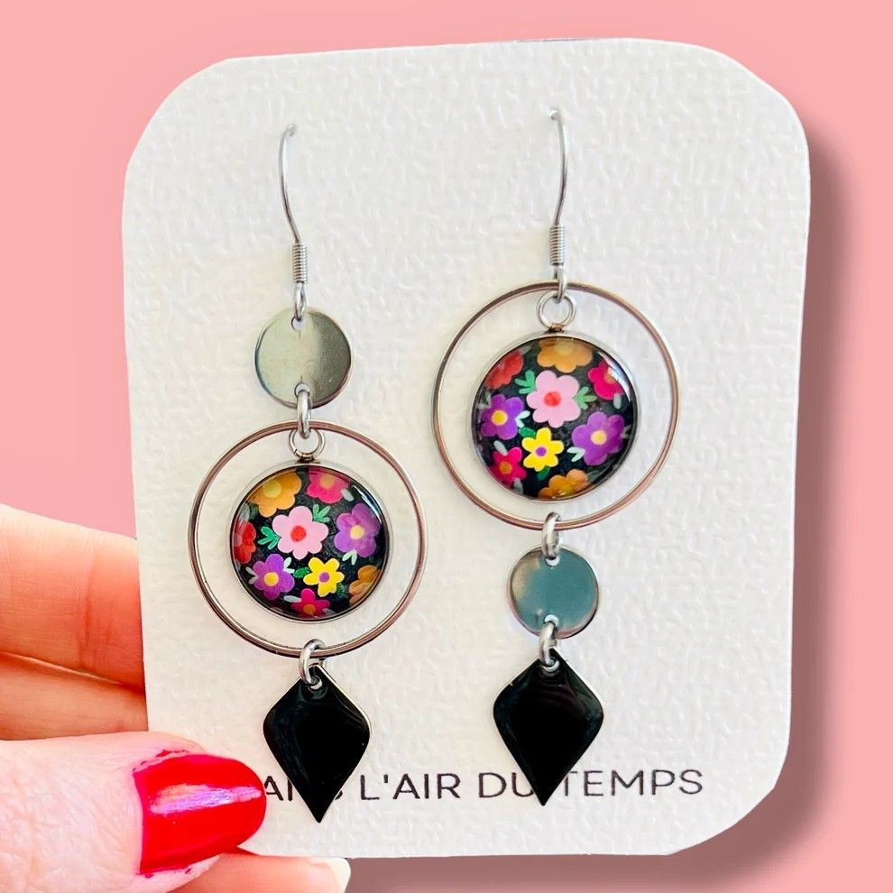 Boucles d'oreilles asymétrique des créatrices "dans l'air du temps" il y'a une attache argentée, avec un rond argenté puis un beau rond coloré fleuri, entouré d'une cercle puis un losange noir tout en bas. L'autre boucle d'oreille a les même éléments mais le rond argenté est en bas du rond fleuri, avant le losange noir. 
