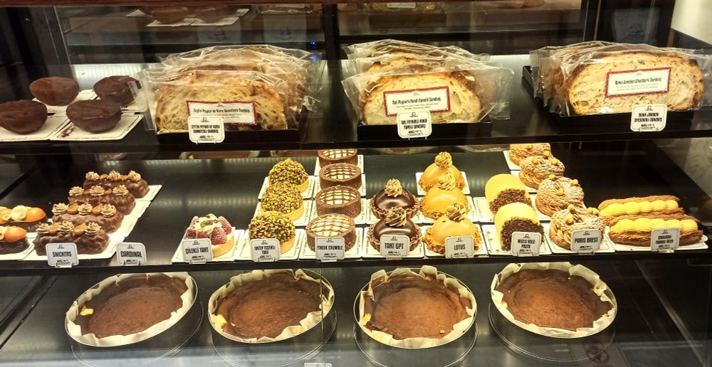 Drei Glasböden voll gestellt mit verschiedenen kleinen und größeren Tortelets und Torten, Eclairs mit gelber Füllung, runde Stücke mit Pistazien umhüllt, andere mit quadratischen Verzierungen aus Zuckerguss oder Kakao, Sahne, Zimt. 