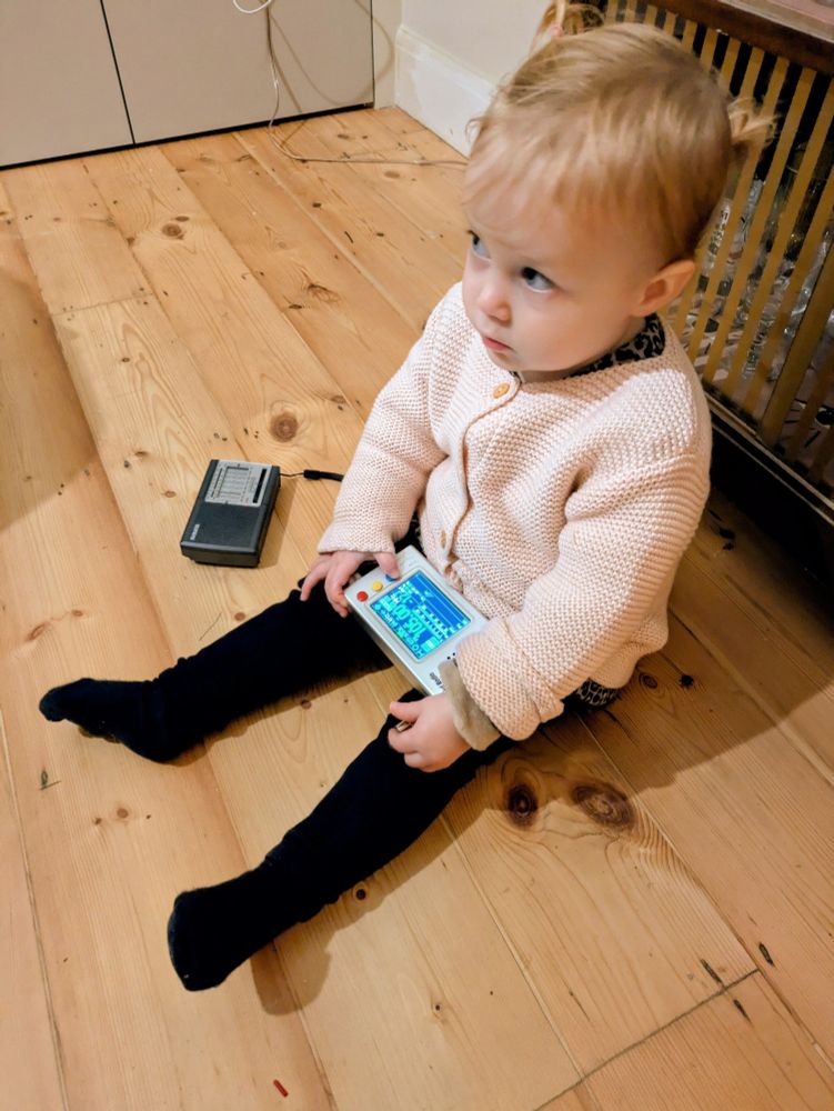 An eighteen month old child is sat on a wooden floor holding one portable radio in her lap while another is on the floor next to her.