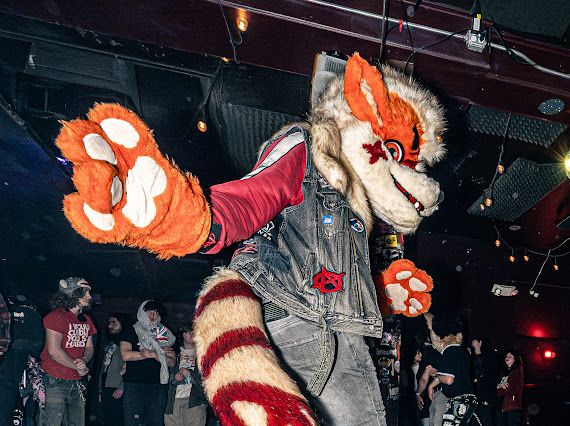 Me in my arcanine fursuit partial with paw outstretched towards the camera. I’m wearing a vest coated in patches and pins. There are many fellow dancers behind me in the photo.