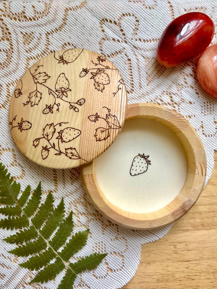Pyrography of strawberry plants on a round wood trinket box displayed on lace with carnelian palmstones and a dried fern