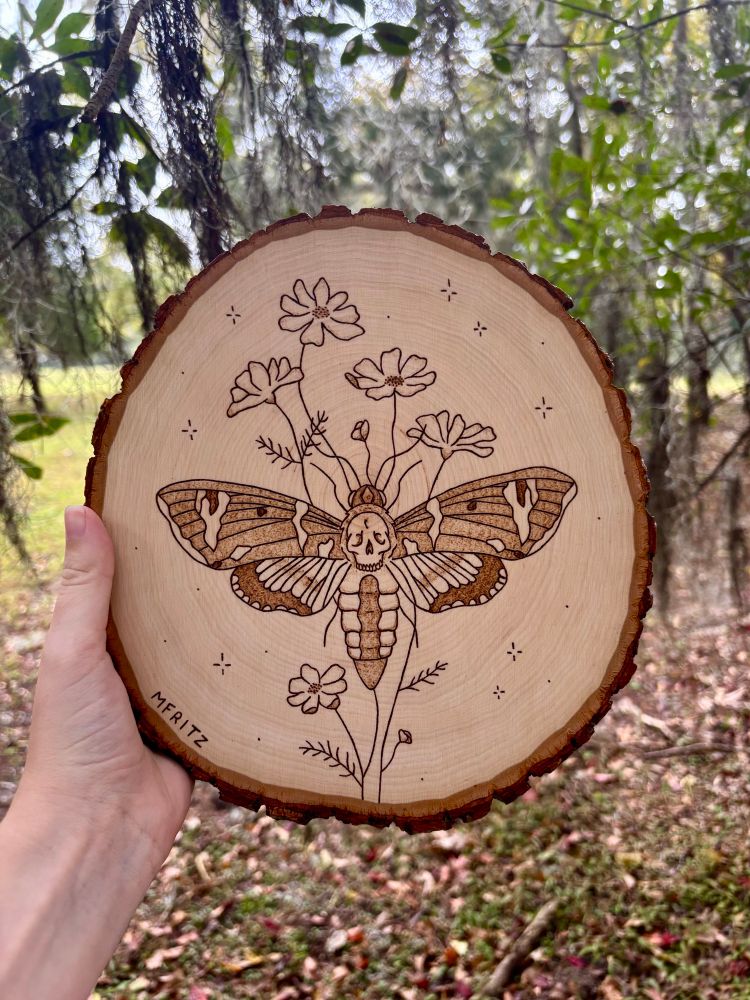 Pyrography of a death’s head hawkmoth with cosmo flowers and sparkles on a wood slice held in front of tree leaves, spanish moss, grass, and fallen autumn leaves