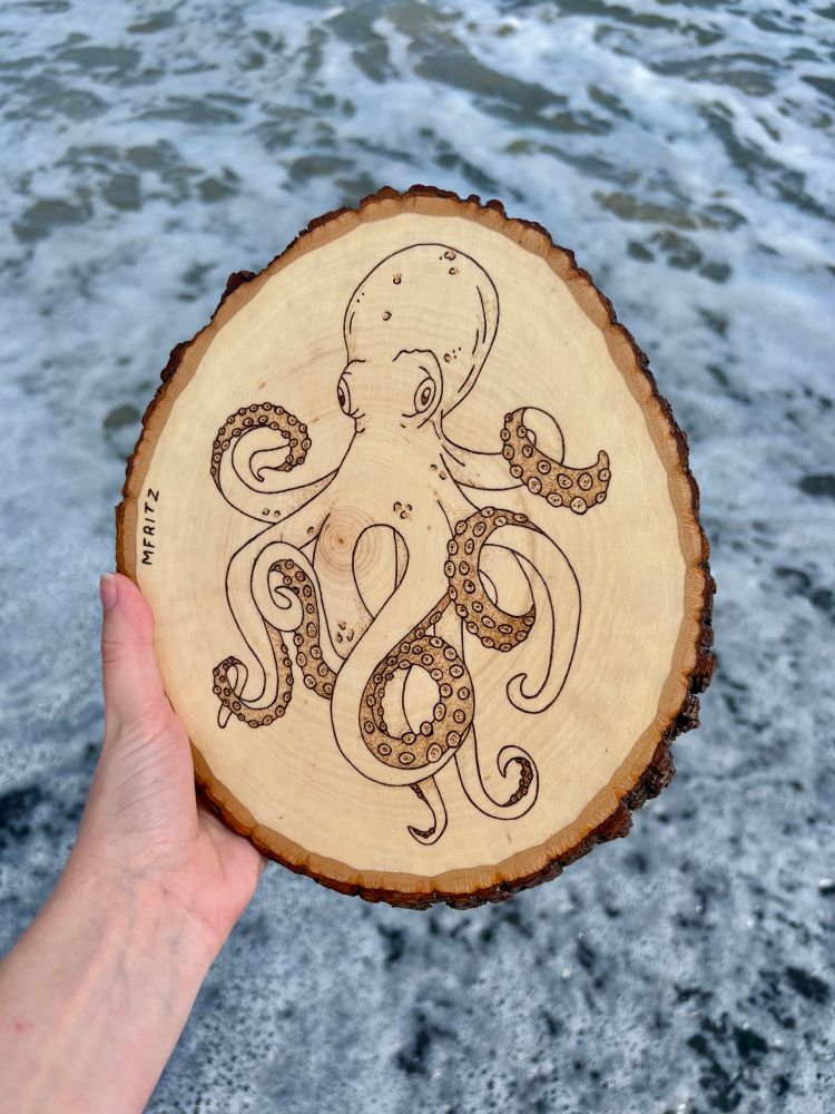 Pyrography of an octopus on a wood slice held in front of the ocean