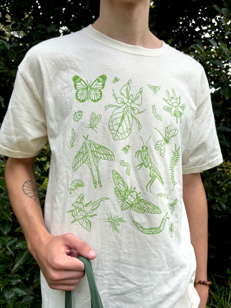 A person standing in front of greenery outside modeling a ivory natural colored tshirt with a light green bug design that includes a butterfly, flies, leaf bug, small moth, caterpillars, stag beetle, ladybug, leopard moth, dragonfly, luna moth, mantis, centipede, isopods, hercules beetle, wasp, cicada, leopard slug, and a bumblebee