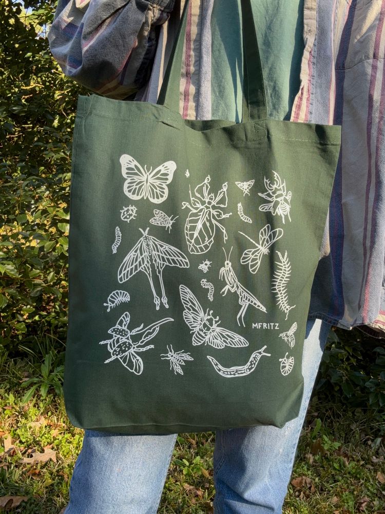 A person wearing a denim striped button down and denim jeans standing in front of greenery outside modeling a forest green tote bag with an off white bug design that includes a butterfly, flies, leaf bug, small moth, caterpillars, stag beetle, ladybug, leopard moth, dragonfly, luna moth, mantis, centipede, isopods, hercules beetle, wasp, cicada, leopard slug, and a bumble bee