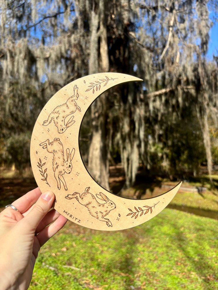 Pyrography of 3 bunnies jumping with ferns and sparkles on a moon shaped wood slice held in front of trees with Spanish moss and grass
