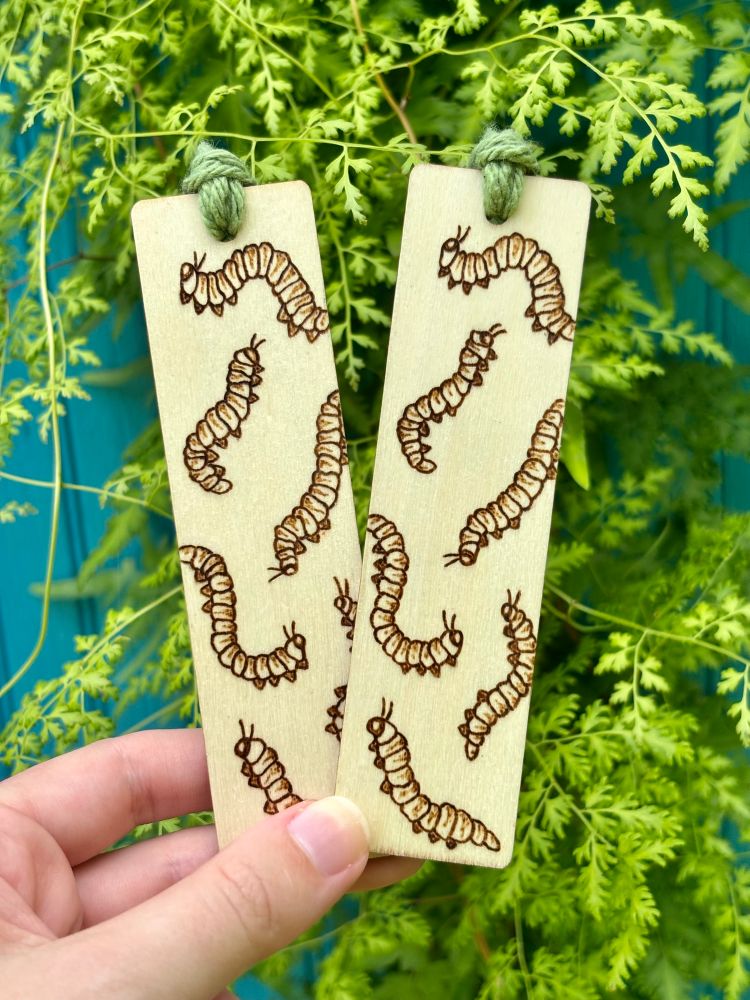 Pyrography of caterpillars on wood bookmarks with green yarn tied to the end and held up in front of ferns