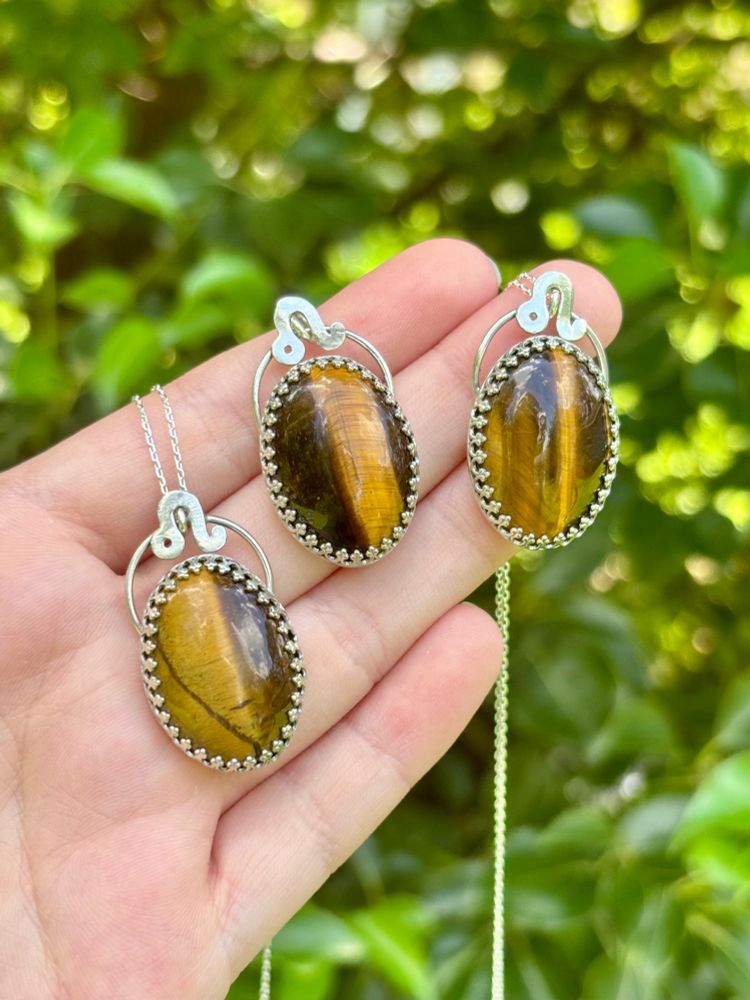 3 tiger’s eye silversmithed necklaces with a Leo symbols held up in front of a leafy green backdrop