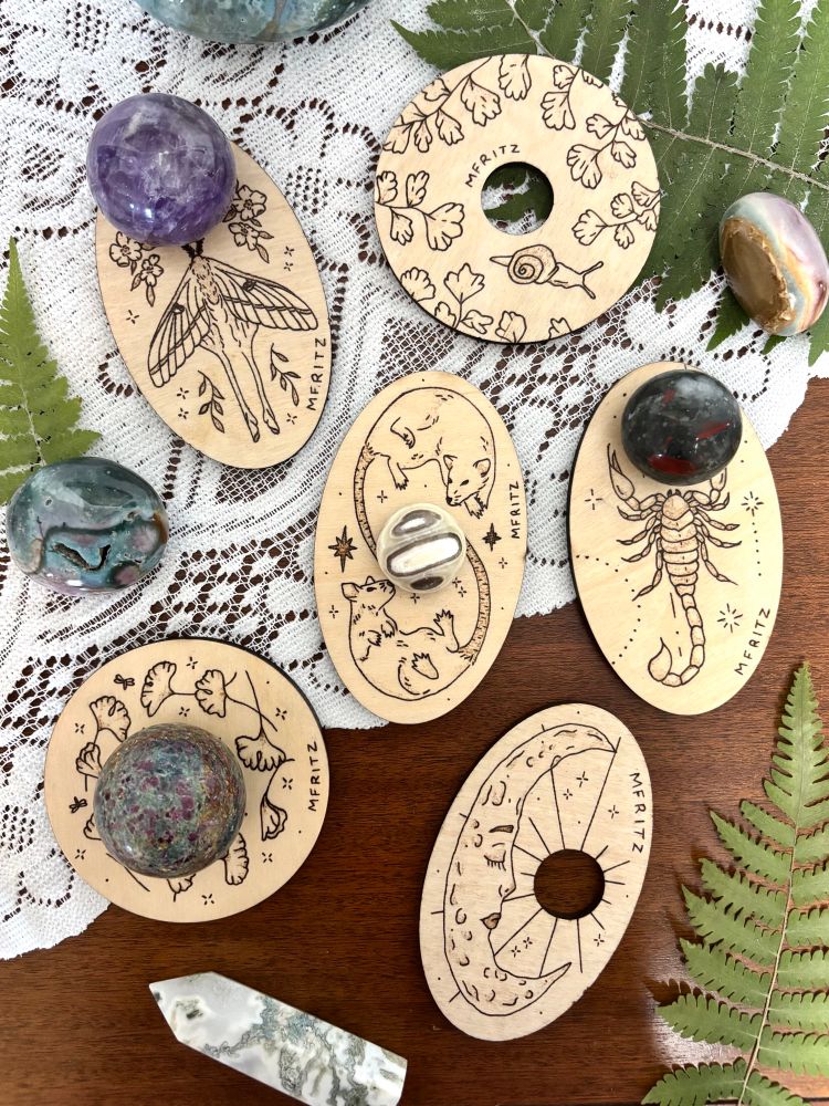 6 wooden crystal sphere stands with pyrography of a luna moth (holding a amethyst sphere), snail and maidenhair ferns, 2 rats (holding a zebra onyx sphere), scorpion (holding a bloodstone sphere), ginkgo leaves (holding a ruby kyanite with fuchsite sphere), and a moon laying on a wood table displayed with lace, dried ferns, moss agate tower, sea jasper palmstone, and a polychrome jasper palmstone.